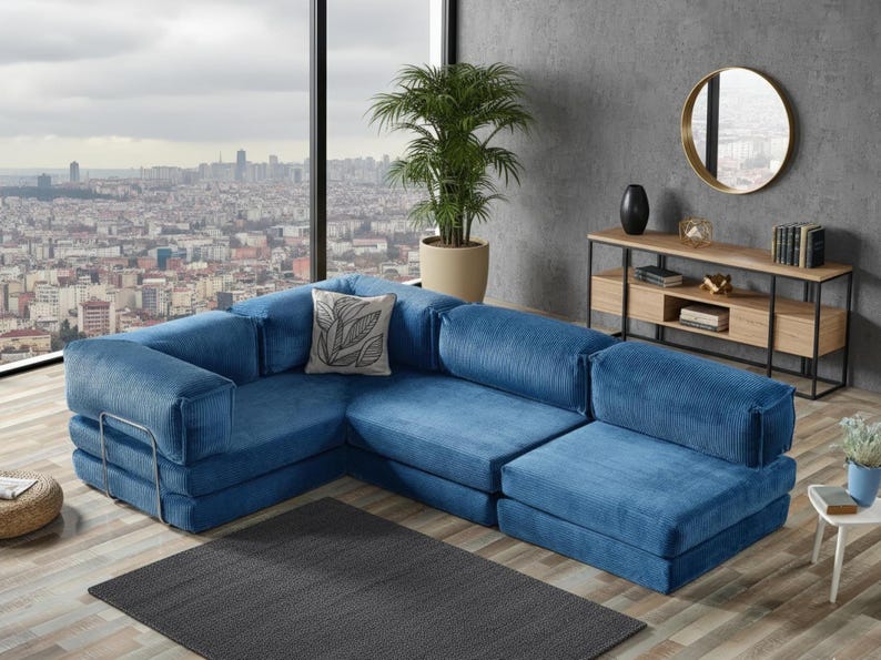 Op de afbeelding: Een grote, modulaire blauwe corduroy hoekbank. De bank heeft een hoekconfiguratie en een decoratief kussen met een bladerenpatroon. De bank staat op een donkergrijs tapijt, met uitzicht op de stad door een groot raam.