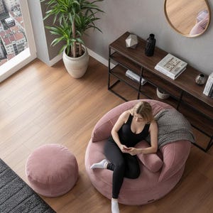 Puede incluir: Un sal&oacute;n moderno con un gran sill&oacute;n puf de pana rosa y un otom&aacute;n a juego. Una mujer est&aacute; sentada en la silla, usando un tel&eacute;fono. Tambi&eacute;n se ve una estanter&iacute;a de madera oscura con decoraci&oacute;n y una gran ventana con vistas a la ciudad.
