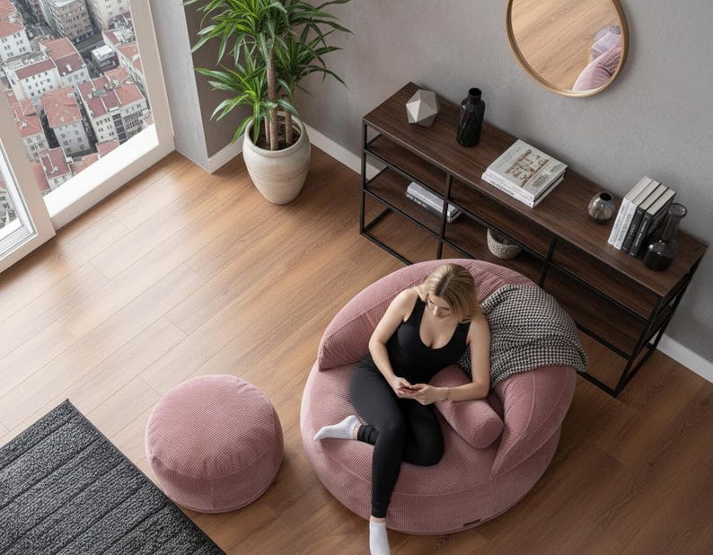Puede incluir: Un sal&oacute;n moderno con un gran sill&oacute;n puf de pana rosa y un otom&aacute;n a juego. Una mujer est&aacute; sentada en la silla, usando un tel&eacute;fono. Tambi&eacute;n se ve una estanter&iacute;a de madera oscura con decoraci&oacute;n y una gran ventana con vistas a la ciudad.