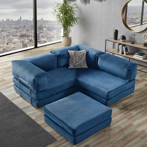 Op de afbeelding: Een blauwe hoekbank van corduroy met een bijpassende voetenbank. De bank heeft een armleuning met kussen en een decoratief kussen. De voetenbank is vierkant en staat voor de bank. De kamer heeft uitzicht op de stad.