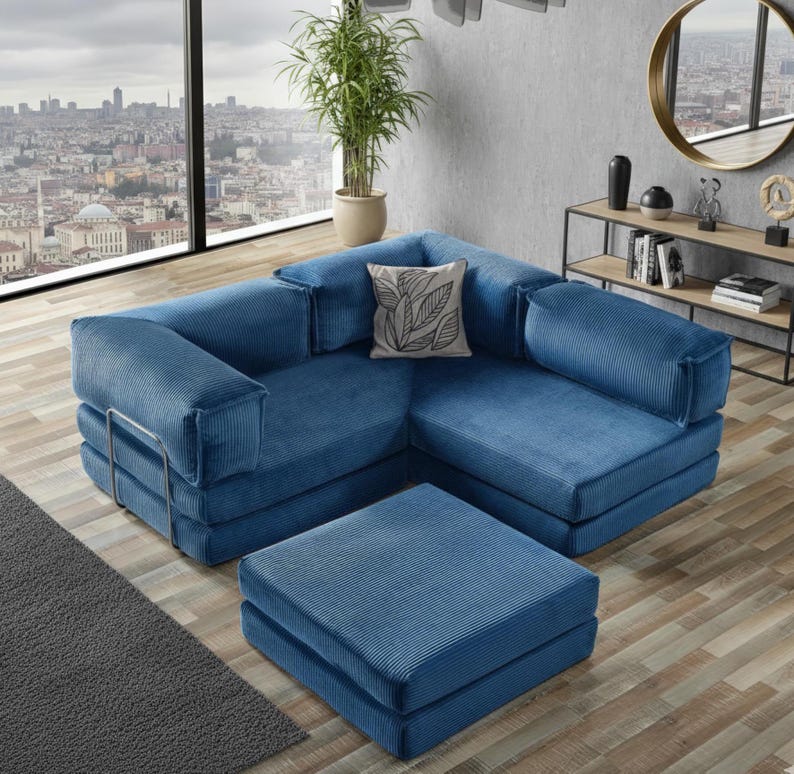 Op de afbeelding: Een blauwe hoekbank van corduroy met een bijpassende voetenbank. De bank heeft een armleuning met kussen en een decoratief kussen. De voetenbank is vierkant en staat voor de bank. De kamer heeft uitzicht op de stad.