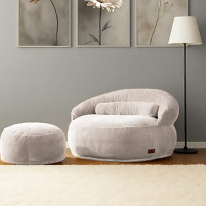 Puede incluir: Un sal&oacute;n acogedor con un sill&oacute;n puf de pana gris claro, un otom&aacute;n a juego y un coj&iacute;n cil&iacute;ndrico. Tres impresiones florales enmarcadas adornan la pared gris. Tambi&eacute;n hay una l&aacute;mpara de pie negra con una pantalla blanca.
