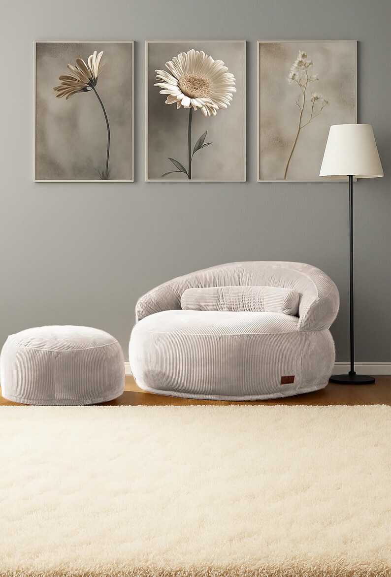 Puede incluir: Un sal&oacute;n acogedor con un sill&oacute;n puf de pana gris claro, un otom&aacute;n a juego y un coj&iacute;n cil&iacute;ndrico. Tres impresiones florales enmarcadas adornan la pared gris. Tambi&eacute;n hay una l&aacute;mpara de pie negra con una pantalla blanca.