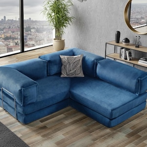 Op de afbeelding: Een moderne, blauwe hoekbank met een corduroy textuur. De L-vormige bank heeft een laag profiel en heeft een decoratief kussen met een bladeren ontwerp. De kamer heeft een groot raam met uitzicht op de stad, een plant en een bijzettafel.