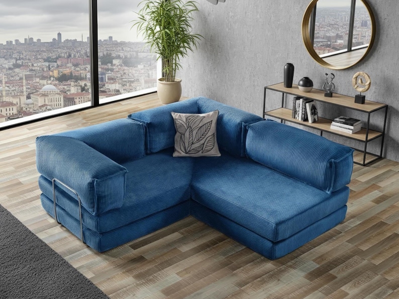 Op de afbeelding: Een moderne, blauwe hoekbank met een corduroy textuur. De L-vormige bank heeft een laag profiel en heeft een decoratief kussen met een bladeren ontwerp. De kamer heeft een groot raam met uitzicht op de stad, een plant en een bijzettafel.