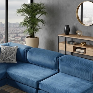 Op de afbeelding: Een hoekbank met blauwe corduroy stof. Een decoratief kussen met een zwart-wit bladmotief ligt op de bank. Een plantenpot en een ronde spiegel zijn op de achtergrond te zien.