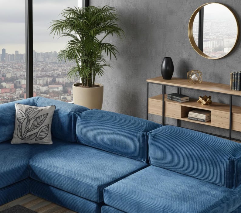 Op de afbeelding: Een hoekbank met blauwe corduroy stof. Een decoratief kussen met een zwart-wit bladmotief ligt op de bank. Een plantenpot en een ronde spiegel zijn op de achtergrond te zien.