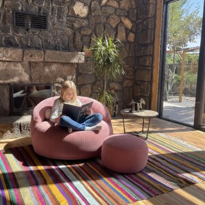 Puede incluir: Escena interior acogedora con un sill&oacute;n de saco de pana rosa y un otom&aacute;n a juego. Una colorida alfombra a rayas y una chimenea de piedra a&ntilde;aden calidez. Una persona lee un libro en la silla.