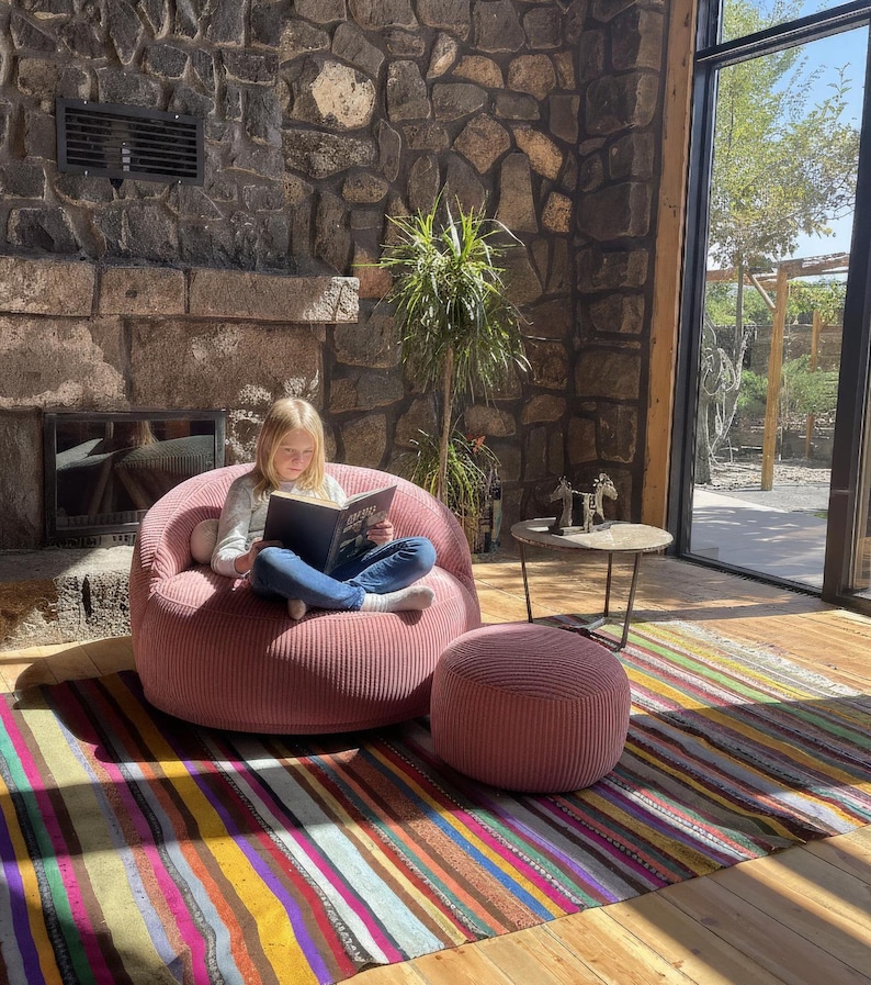 Puede incluir: Escena interior acogedora con un sill&oacute;n de saco de pana rosa y un otom&aacute;n a juego. Una colorida alfombra a rayas y una chimenea de piedra a&ntilde;aden calidez. Una persona lee un libro en la silla.