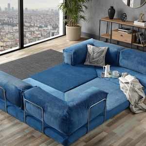 Op de afbeelding: Een modulaire blauwe corduroy bank met metalen armleuningen. De bank is opgesteld in een hoekconfiguratie, met een decoratief kussen, een dienblad met een theepot en kopjes en een opgevouwen deken. Op de achtergrond is een stadsgezicht te zien.