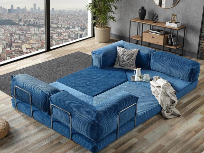 Op de afbeelding: Een modulaire blauwe corduroy bank met metalen armleuningen. De bank is opgesteld in een hoekconfiguratie, met een decoratief kussen, een dienblad met een theepot en kopjes en een opgevouwen deken. Op de achtergrond is een stadsgezicht te zien.