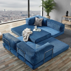 Op de afbeelding: Modulaire blauwe corduroy bank met metalen armleuningen. De hoekbank is opgesteld in een hoekconfiguratie, met een vierkante poef en een rechthoekig tapijt. Een skyline is zichtbaar door een groot raam.