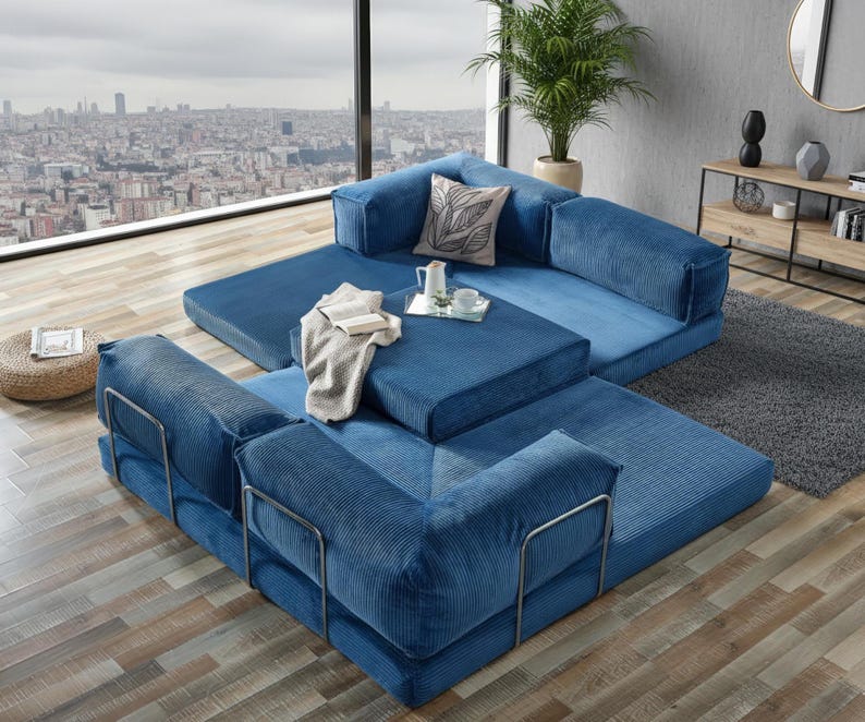 Op de afbeelding: Modulaire blauwe corduroy bank met metalen armleuningen. De hoekbank is opgesteld in een hoekconfiguratie, met een vierkante poef en een rechthoekig tapijt. Een skyline is zichtbaar door een groot raam.