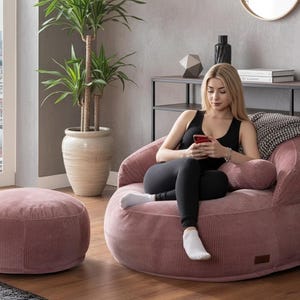 Puede incluir: Un sill&oacute;n de pana rosa empolvado y un otom&aacute;n a juego. El sill&oacute;n tiene forma redondeada con respaldo bajo y reposabrazos. El otom&aacute;n es redondo y bajo. Una mujer est&aacute; sentada en el sill&oacute;n.