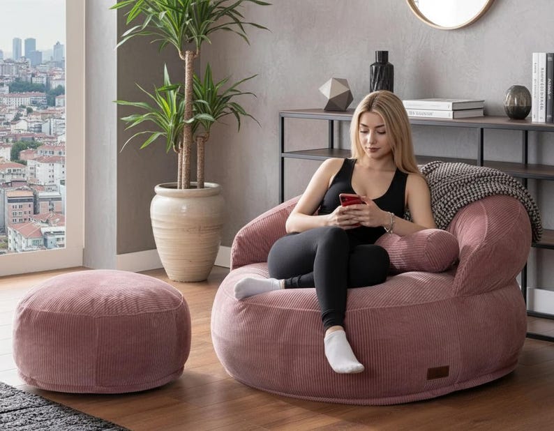 Puede incluir: Un sill&oacute;n de pana rosa empolvado y un otom&aacute;n a juego. El sill&oacute;n tiene forma redondeada con respaldo bajo y reposabrazos. El otom&aacute;n es redondo y bajo. Una mujer est&aacute; sentada en el sill&oacute;n.