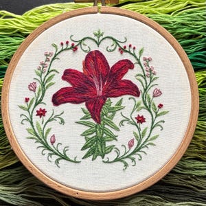May include: A circular wooden embroidery hoop containing white fabric with a hand-stitched design of a large, deep red lily centered within a delicate, circular wreath of green vines, small pink flower buds, and tiny red star-shaped blossoms.