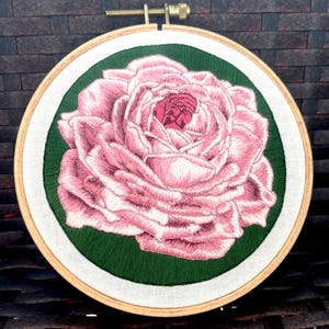 May include: An embroidered pink rose in a wooden hoop. The rose is set against a green backdrop, with a white border. The embroidery is held in place with a brass screw and a wooden frame.