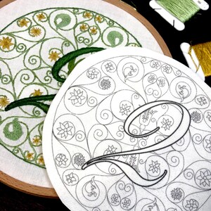 May include: A wooden embroidery hoop containing white fabric with a partially stitched green and yellow floral pattern, alongside a separate white fabric piece featuring a printed black outline of the letter Q surrounded by intricate floral motifs.
