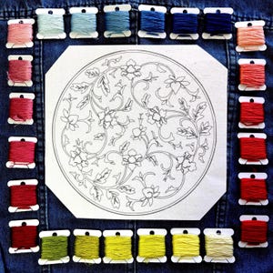 May include: A selection of embroidery threads in various colours, encircling a floral design printed on paper. The thread colours include pinks, blues, greens, and yellows. The background is a denim fabric.