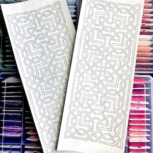 May include: Two rectangular pieces of white fabric with intricate black geometric patterns. The designs are reminiscent of Celtic knotwork. The background shows organized spools of colorful embroidery floss, suggesting a craft or textile project.