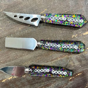 Puede incluir: Tres cuchillos de queso con hojas plateadas y mangos brillantes. Los mangos están decorados con purpurina verde, morada y dorada y deletrean "MARDI PARDI" en letras blancas. Los cuchillos están sobre una superficie de madera.