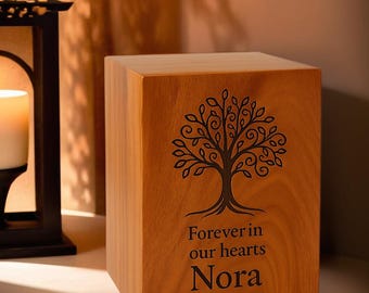 May include: A wooden urn with a tree design and the inscription "Forever in our hearts Nora 2008-2021." The urn is a light brown color and has a square shape. A lit candle and a decorative lantern are in the background.