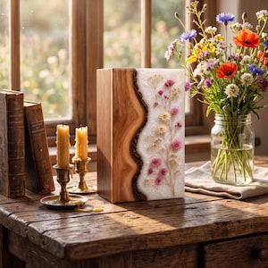 May include: A rectangular wooden urn featuring a natural live edge on one side, seamlessly joined to a white resin panel embedded with delicate pink and cream dried flowers, standing upright on a rustic wooden surface next to two candles.