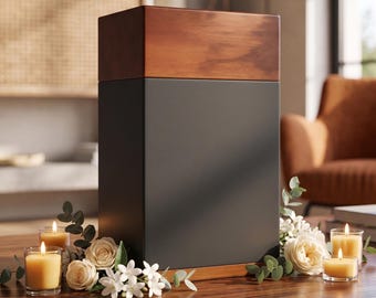 May include: A modern, rectangular urn with a dark gray body and a wooden top and base. The urn is decorated with white roses, small white flowers, and green leaves. Lit candles in glass holders are placed around the urn.