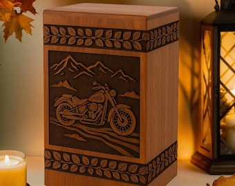 Könnte beinhalten: Eine Holzurne mit einem geschnitzten Motorrad-Design. Die rechteckige Urne besteht aus hellbraunem Holz mit einer dunkleren braunen, geschnitzten Tafel mit einem Motorrad und einer Berglandschaft. Blattförmige Ränder umrahmen die geschnitzte Tafel und die Oberseite der Urne.