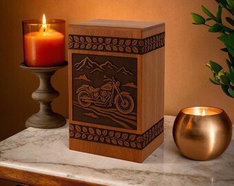 Könnte beinhalten: Eine Holzurne mit einem geschnitzten Motorrad-Design und einer Berglandschaft. Die rechteckige Urne hat Blattbordüren. Eine brennende Kerze in einem Glashalter und eine goldene Kerze in einem runden Halter befinden sich auf beiden Seiten.