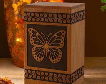 Puede incluir: Una urna de madera con diseño de mariposa. La urna rectangular es de madera marrón claro, con un panel oscuro con una mariposa recortada. Patrones decorativos de hojas bordean la parte superior e inferior.