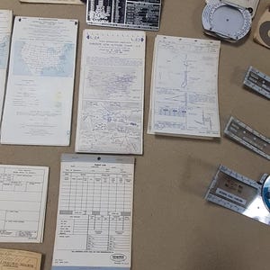May include: A collection of vintage aviation navigation tools and documents, including a knee board, flight computer, charts, and a flight log. The items are arranged on a neutral-toned surface.