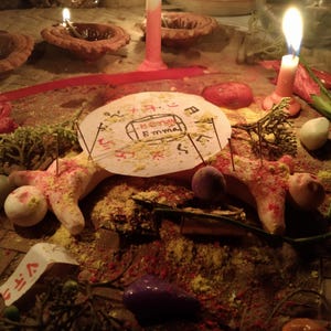 Puede incluir: Un disco de papel circular con el nombre Emma escrito en el centro, rodeado de s&iacute;mbolos rojos y negros. El disco est&aacute; colocado sobre una figura de masa clara con agujas insertadas, rodeado de polvo amarillo y velas encendidas.