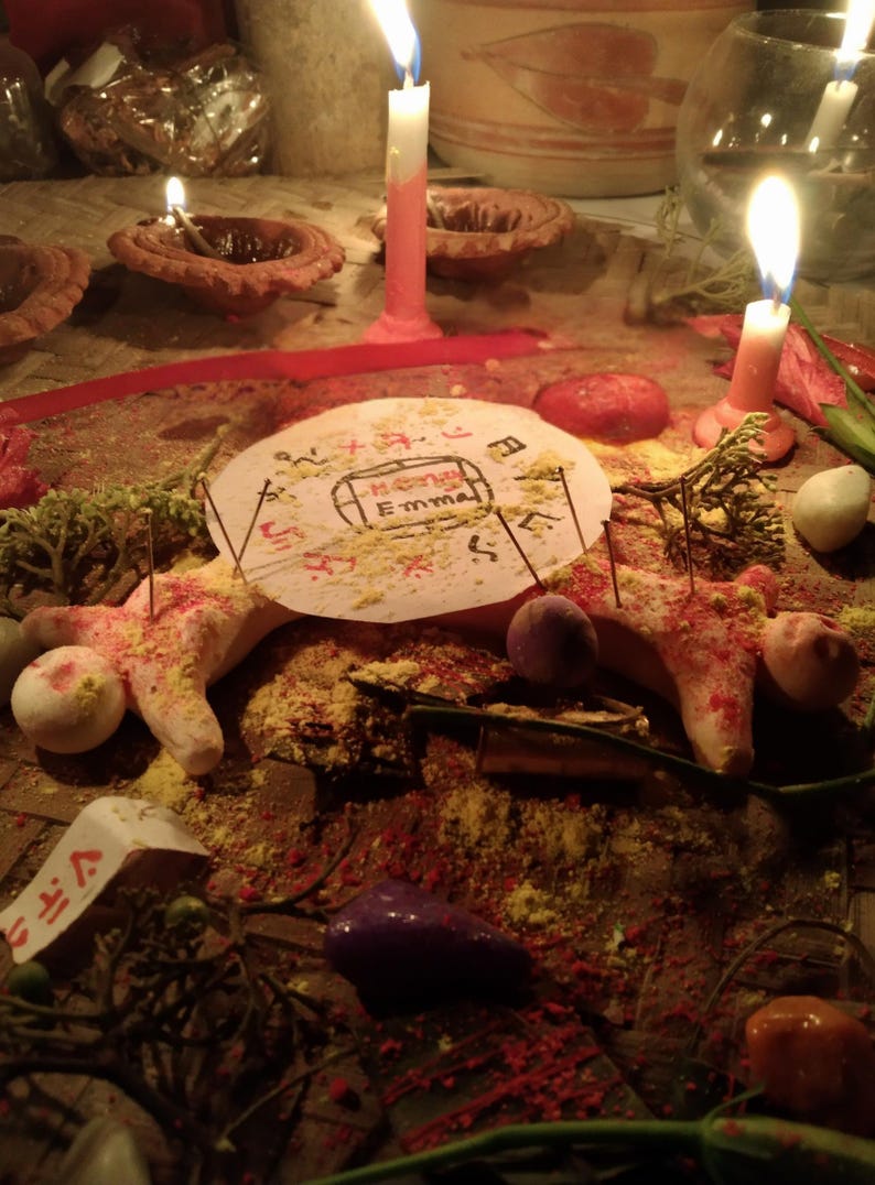 Puede incluir: Un disco de papel circular con el nombre Emma escrito en el centro, rodeado de s&iacute;mbolos rojos y negros. El disco est&aacute; colocado sobre una figura de masa clara con agujas insertadas, rodeado de polvo amarillo y velas encendidas.