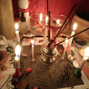 May include: A circular paper mat featuring a complex occult-style geometric design with symbols and text, surrounded by multiple lit white candles, black incense sticks in a silver-toned metal holder, and scattered small colorful stones.