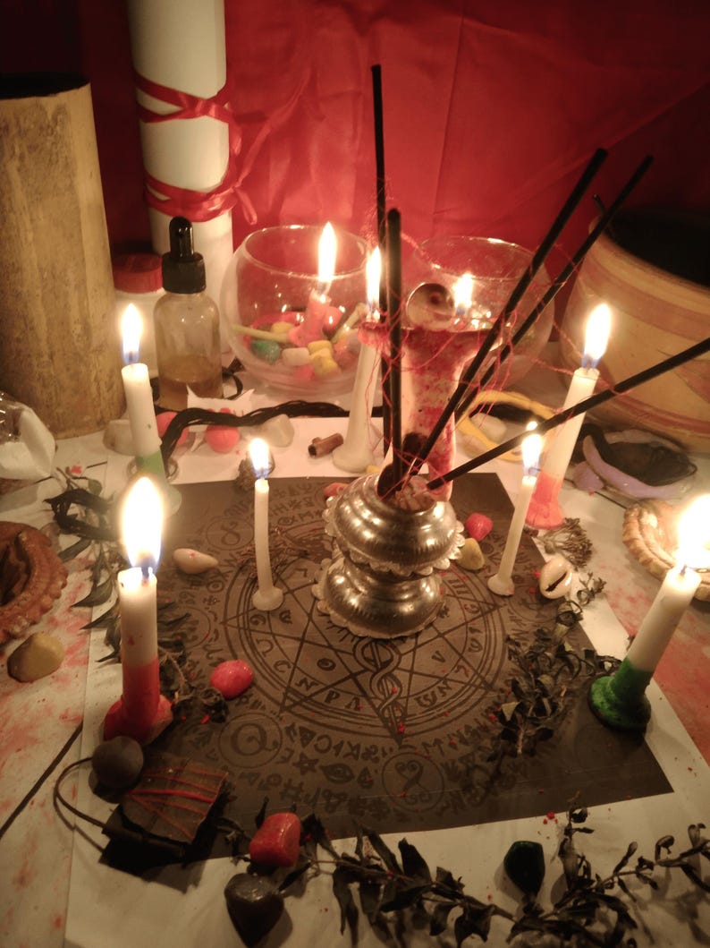 May include: A circular paper mat featuring a complex occult-style geometric design with symbols and text, surrounded by multiple lit white candles, black incense sticks in a silver-toned metal holder, and scattered small colorful stones.
