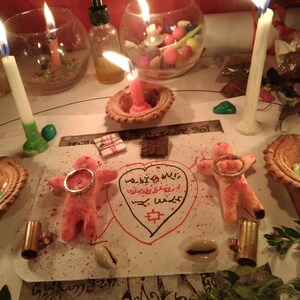 May include: A spiritual arrangement featuring lit candles, small glass bowls, and a central drawing with symbols and text. Two pink figures with rings are positioned on the paper, alongside shells and other ritualistic items. The scene is illuminated by candlelight.