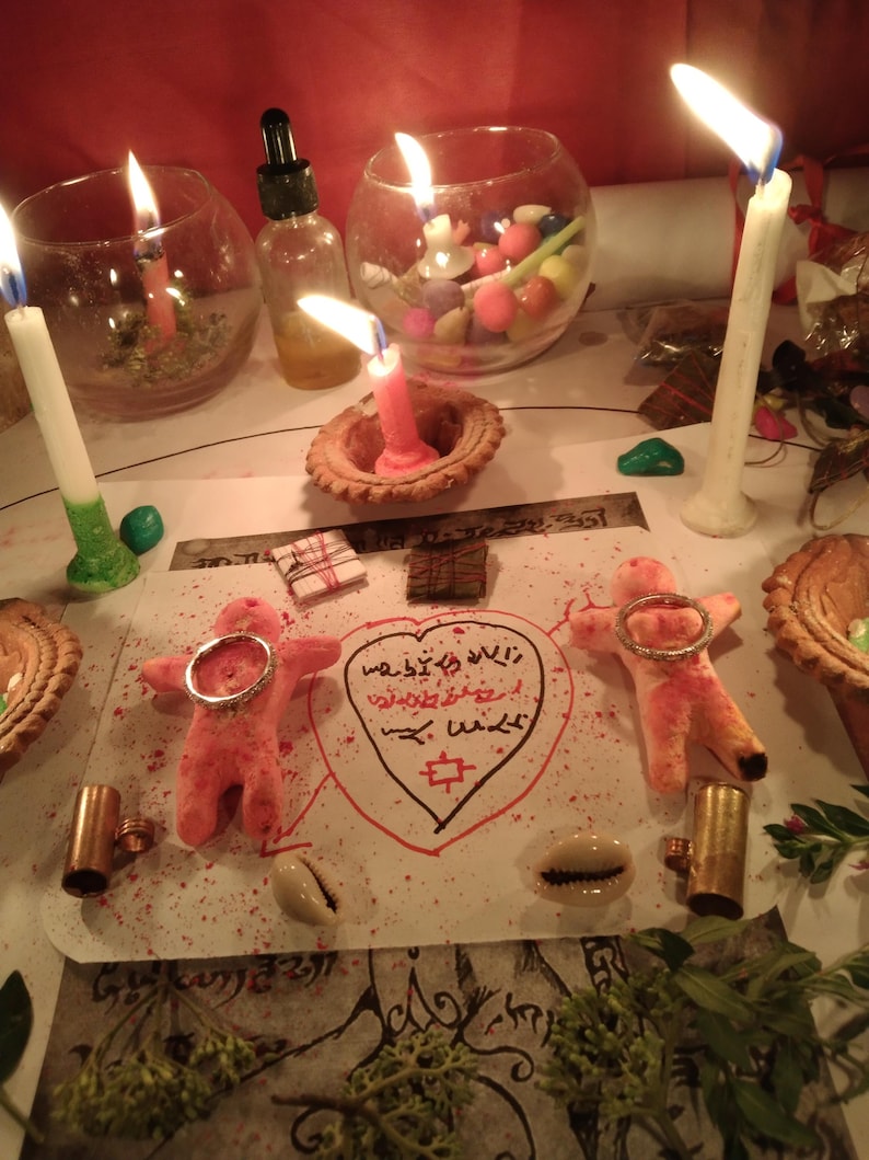 May include: A spiritual arrangement featuring lit candles, small glass bowls, and a central drawing with symbols and text. Two pink figures with rings are positioned on the paper, alongside shells and other ritualistic items. The scene is illuminated by candlelight.