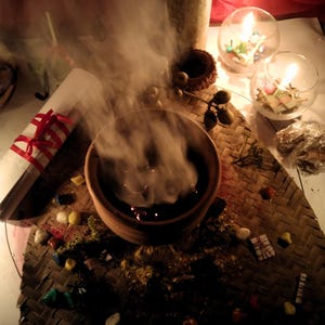 Puede incluir: Un cuenco de barro marr&oacute;n que emite un espeso humo blanco, colocado sobre una esterilla tejida con piedras de colores dispersas, un papel blanco enrollado atado con una cinta roja, dos cuencos de cristal con velas encendidas y una vela cil&iacute;ndrica alta.