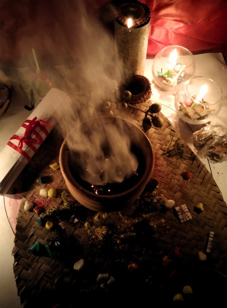 Puede incluir: Un cuenco de barro marr&oacute;n que emite un espeso humo blanco, colocado sobre una esterilla tejida con piedras de colores dispersas, un papel blanco enrollado atado con una cinta roja, dos cuencos de cristal con velas encendidas y una vela cil&iacute;ndrica alta.