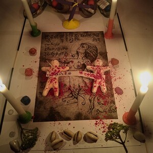 Puede incluir: Una impresi&oacute;n rectangular con una ilustraci&oacute;n central de una figura rodeada de escritura, dos peque&ntilde;as figuras humanoides de color beige con motas rojas, cuatro velas encendidas, conchas de cauri dispersas, piedras de colores y ramitas verdes.