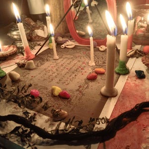 May include: A rectangular paper with printed text and illustrations is surrounded by several lit white candles, small colorful stones, and dried plant stems. A red-framed mirror stands behind the paper, reflecting the candlelight and surroundings.
