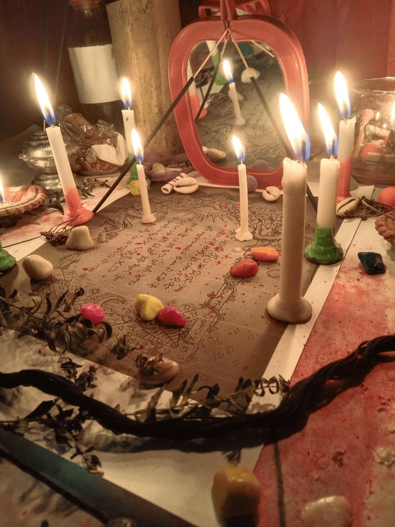 May include: A rectangular paper with printed text and illustrations is surrounded by several lit white candles, small colorful stones, and dried plant stems. A red-framed mirror stands behind the paper, reflecting the candlelight and surroundings.
