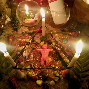 Puede incluir: Un arreglo ritual con una figura humana central de arcilla rosa rodeada por un tri&aacute;ngulo de tres velas blancas encendidas. La escena incluye piedras coloridas dispersas, peque&ntilde;os anillos de metal, cuadrados de chocolate oscuro y un pergamino blanco.