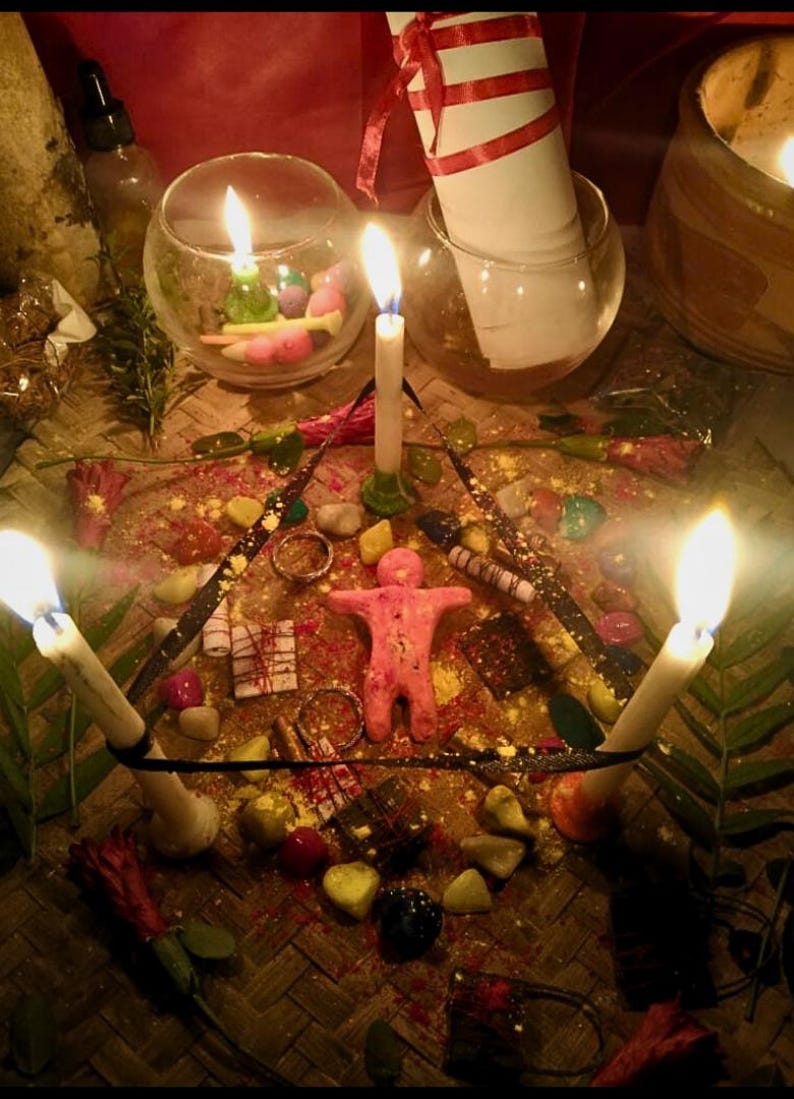 Puede incluir: Un arreglo ritual con una figura humana central de arcilla rosa rodeada por un tri&aacute;ngulo de tres velas blancas encendidas. La escena incluye piedras coloridas dispersas, peque&ntilde;os anillos de metal, cuadrados de chocolate oscuro y un pergamino blanco.