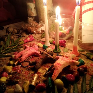 May include: Three small clay figures in pink, brown, and white are arranged on a woven mat surrounded by colorful stones, green leaves, and red flowers. Three tall white candles with lit flames stand behind the figures, with a glass bowl nearby.
