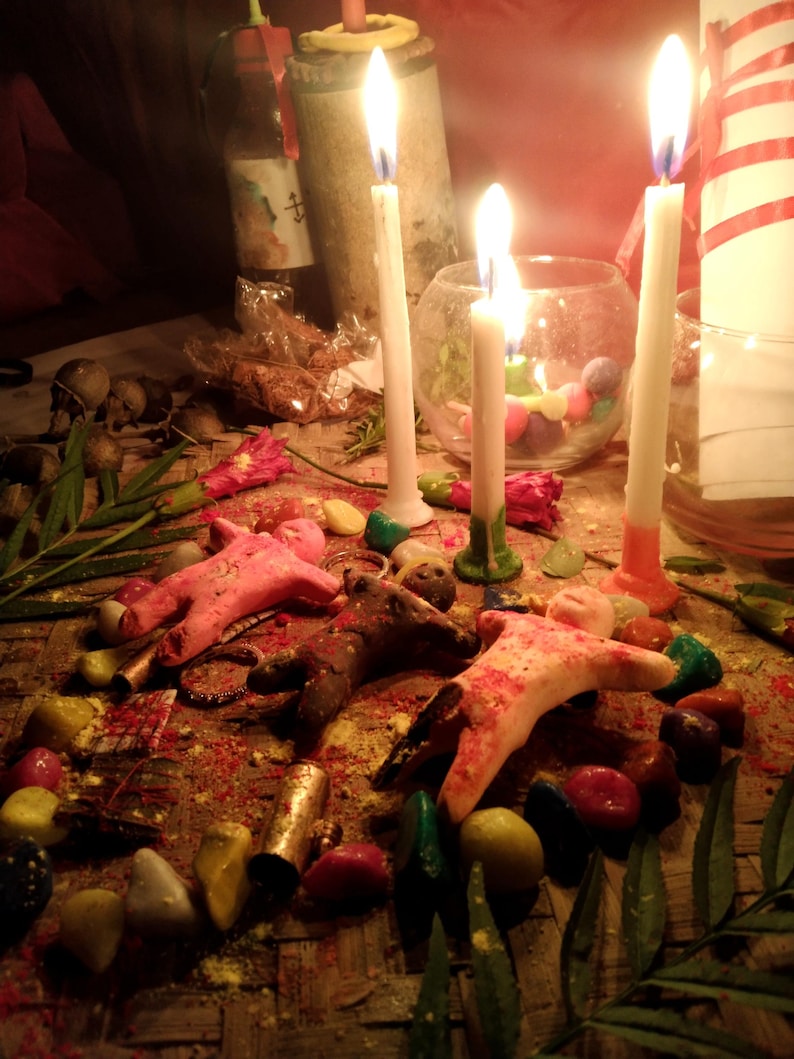 May include: Three small clay figures in pink, brown, and white are arranged on a woven mat surrounded by colorful stones, green leaves, and red flowers. Three tall white candles with lit flames stand behind the figures, with a glass bowl nearby.