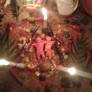 May include: Two pink human-shaped wax figures stand together at the center of a triangular arrangement of three lit white candles, colorful stones, small wrapped packages, and red hibiscus flowers on a woven mat with glass candle holders.