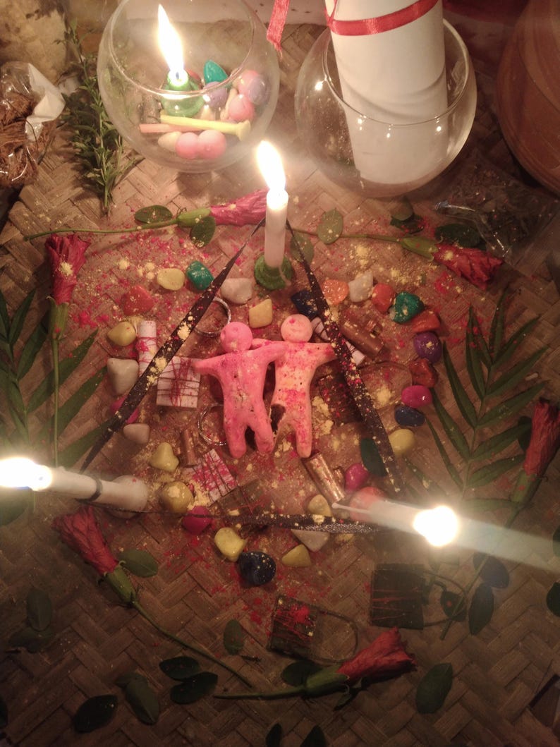 May include: Two pink human-shaped wax figures stand together at the center of a triangular arrangement of three lit white candles, colorful stones, small wrapped packages, and red hibiscus flowers on a woven mat with glass candle holders.