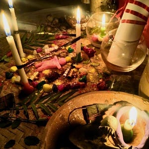 Puede incluir: Una colecci&oacute;n de objetos rituales que incluye varias velas blancas encendidas, una peque&ntilde;a figura rosa, piedras de colores, hojas verdes y un pergamino blanco enrollado con una cinta roja, dispuestos sobre una estera tejida con un cuenco de barro.
